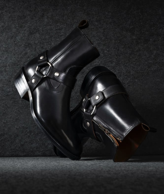 An angled view of a pair of black leather moto boots with strap details and a side zip closure.  
