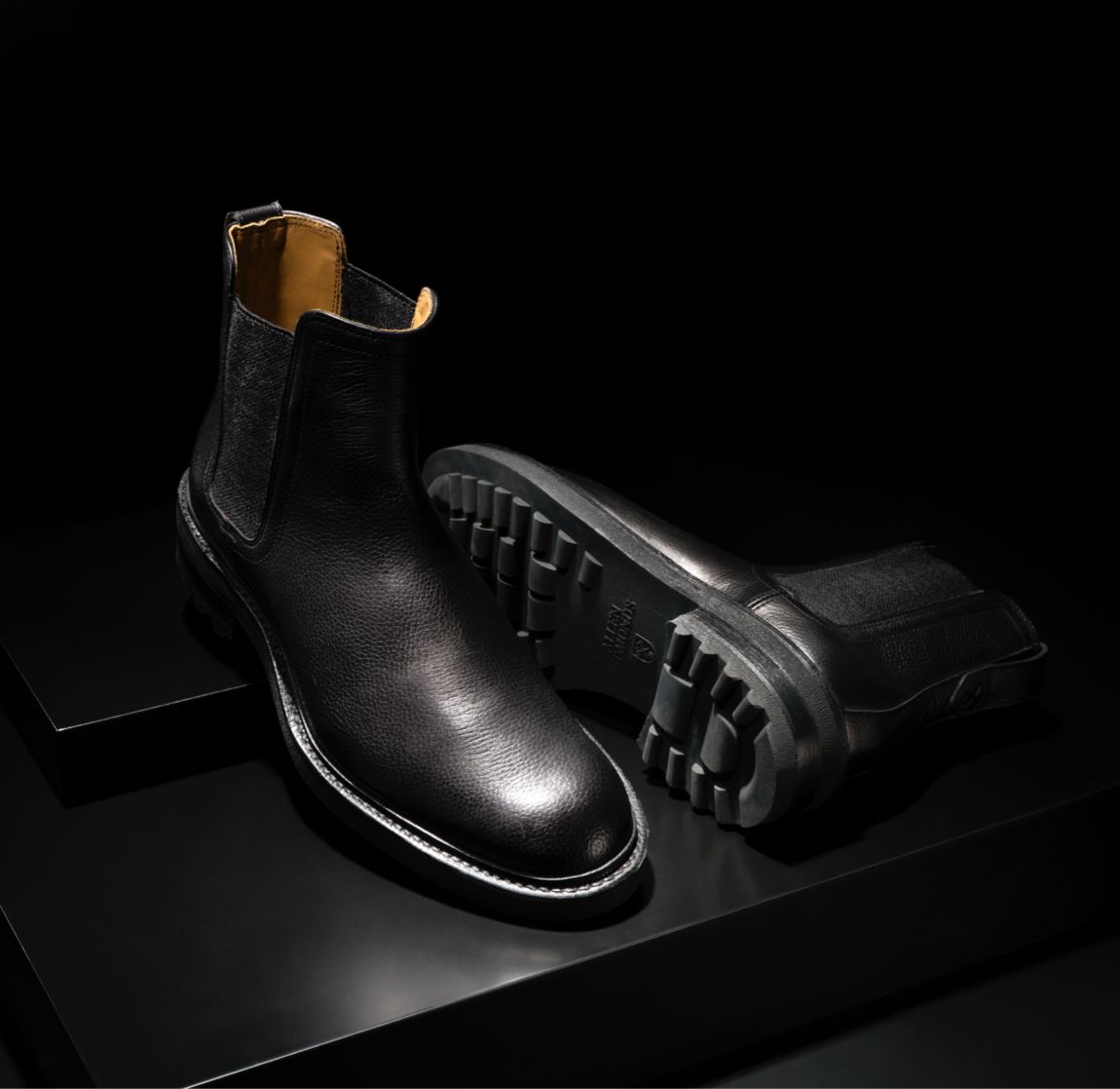 A pair of men’s Chelsea boots in black leather, with one boot laying on its side.