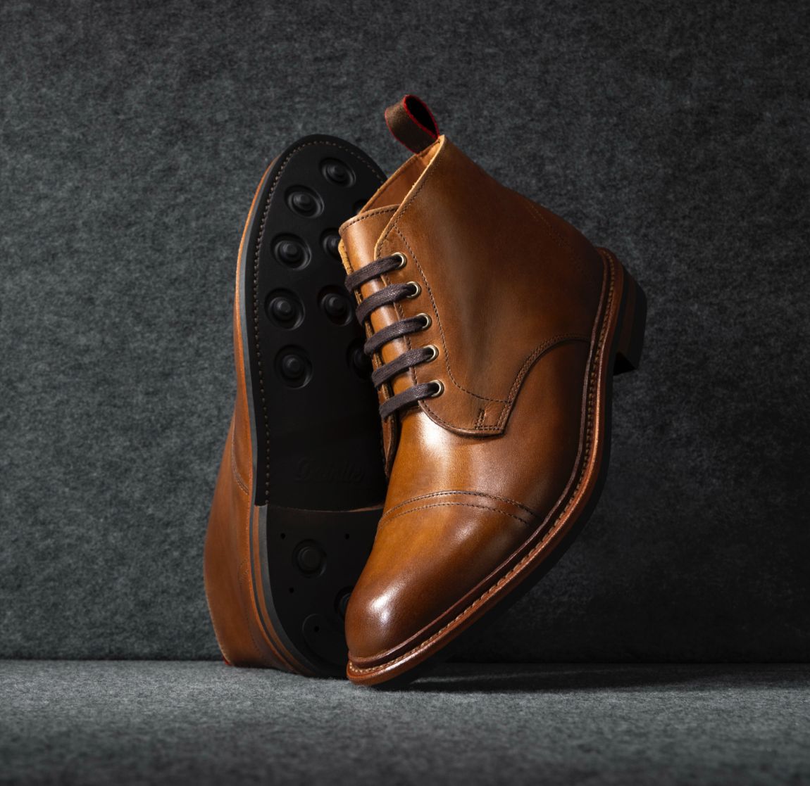 A pair of men’s brown cap-toe boots displayed vertically with a view of the Dainite rubber sole.   