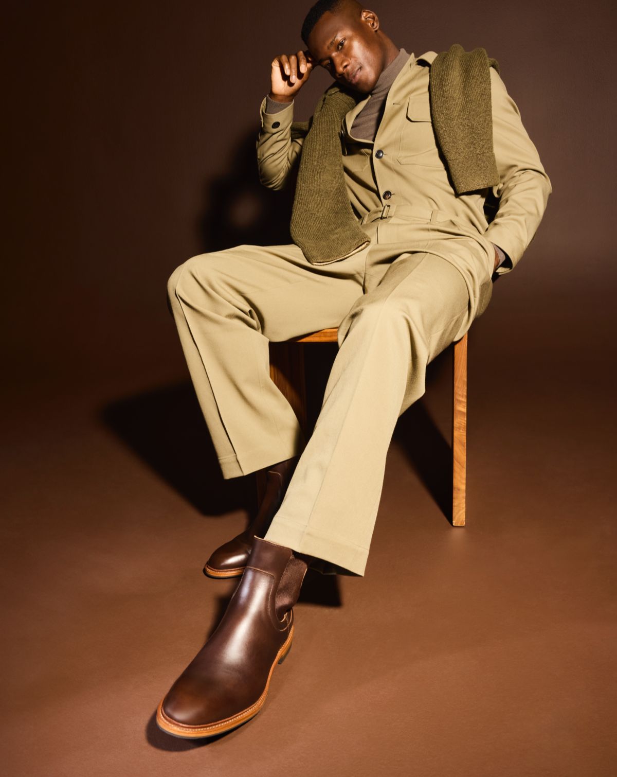 A man sitting in a chair modeling a tan pair of pants and matching jacket, with a green sweater draped over his shoulder and a pair of brown leather Chelsea boots.  