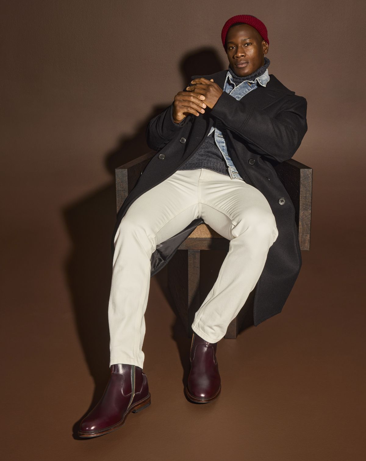 A man sitting in a chair modeling off-white jeans, a black trench coat, denim jacket, and a pair of burgundy zip-up boots.  