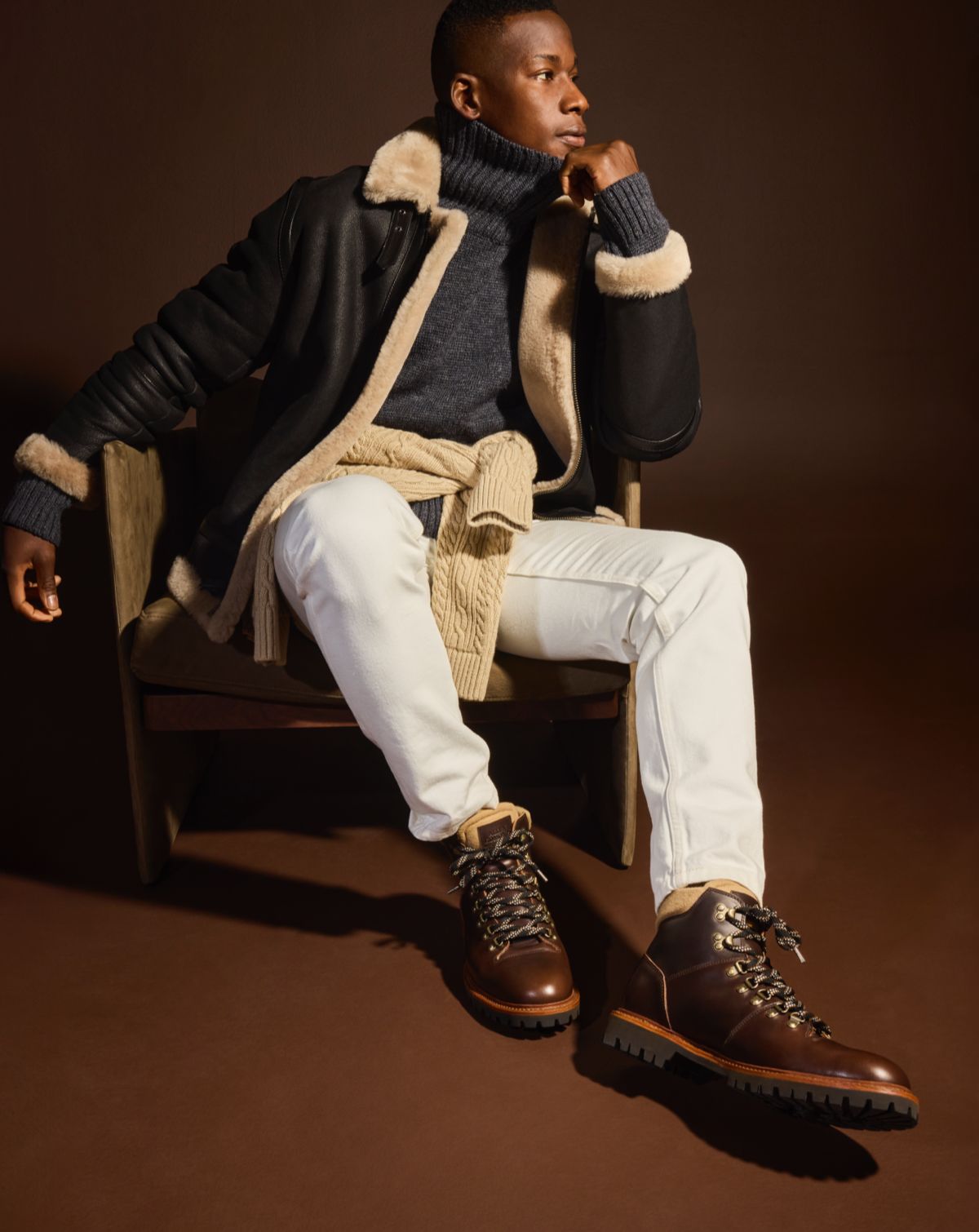 A man sitting in a chair modeling off-white jeans, a gray turtleneck sweater, a black bomber jacket, and a pair of brown hiker boots.  