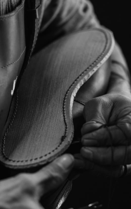 A pair of hands holding a men’s dress shoe with the sole facing up. A needle and thread are being used to make the final stitches. 