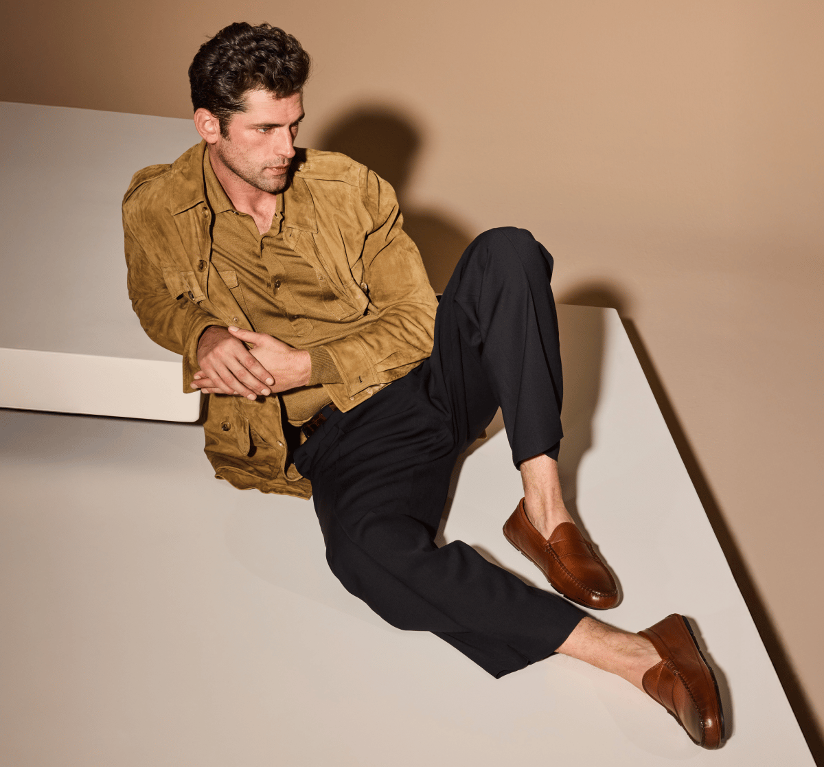 Man seated on white surface wearing Chili Brown Leather Apex Driving Loafer, styled with black trousers and a tan suede jacket 