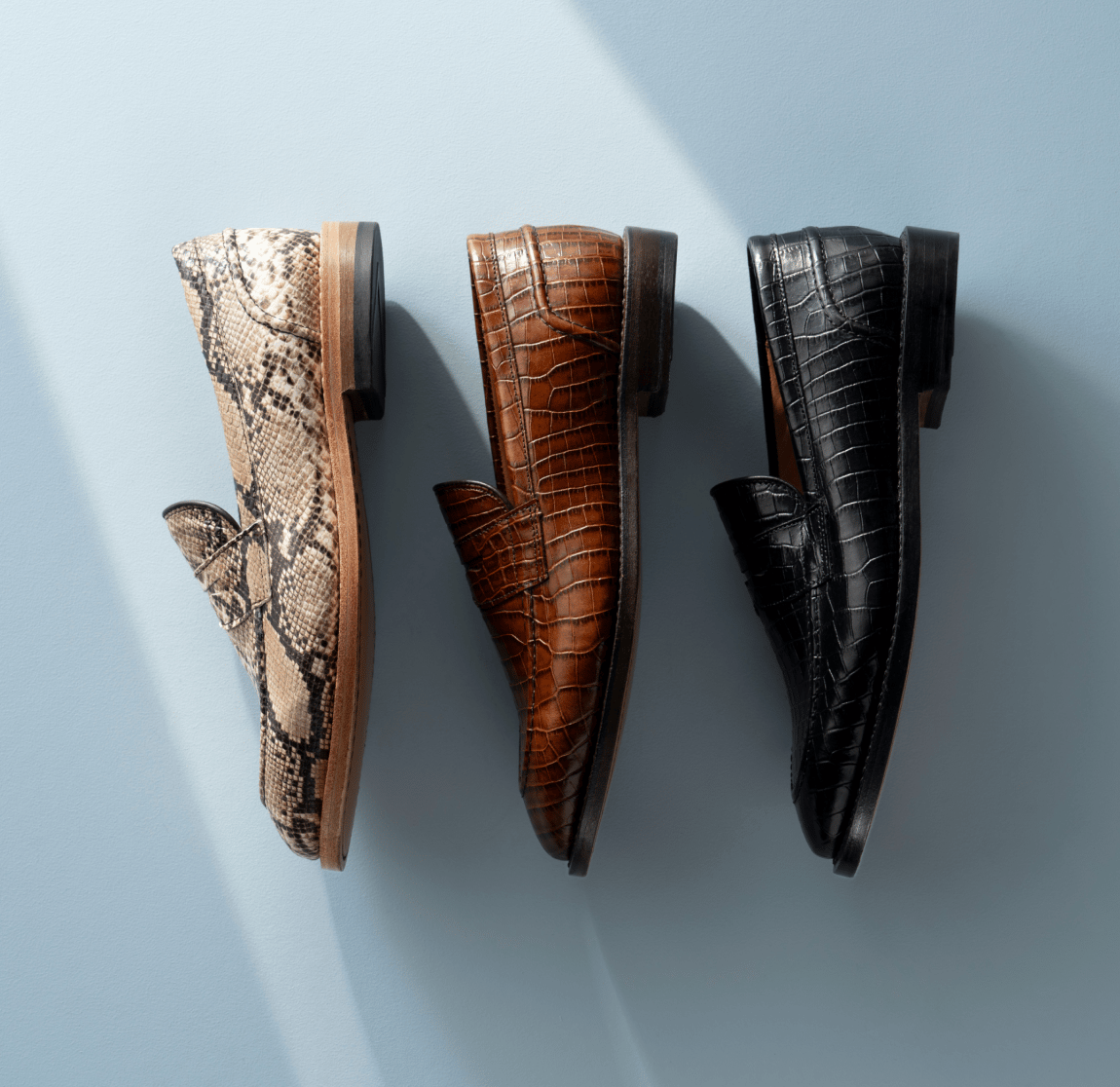 Allen Edmonds Randolph 2.0 Penny Loafers displayed in python leather, brown croco leather, and black croco leather against a light blue background