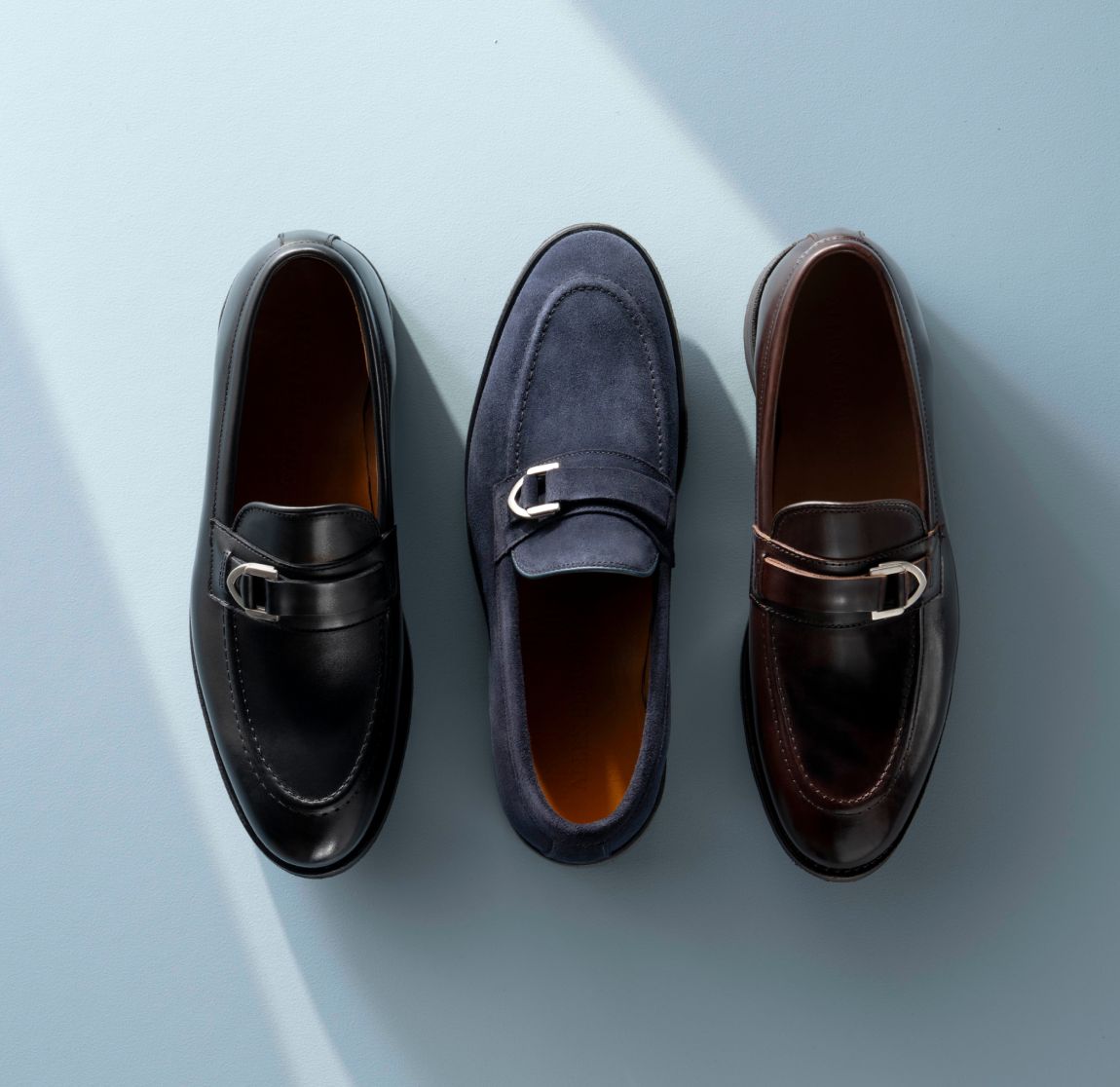 Dark brown, black, and navy leather Allen Edmonds Rollins buckle loafers with silver buckles against a light blue background. 