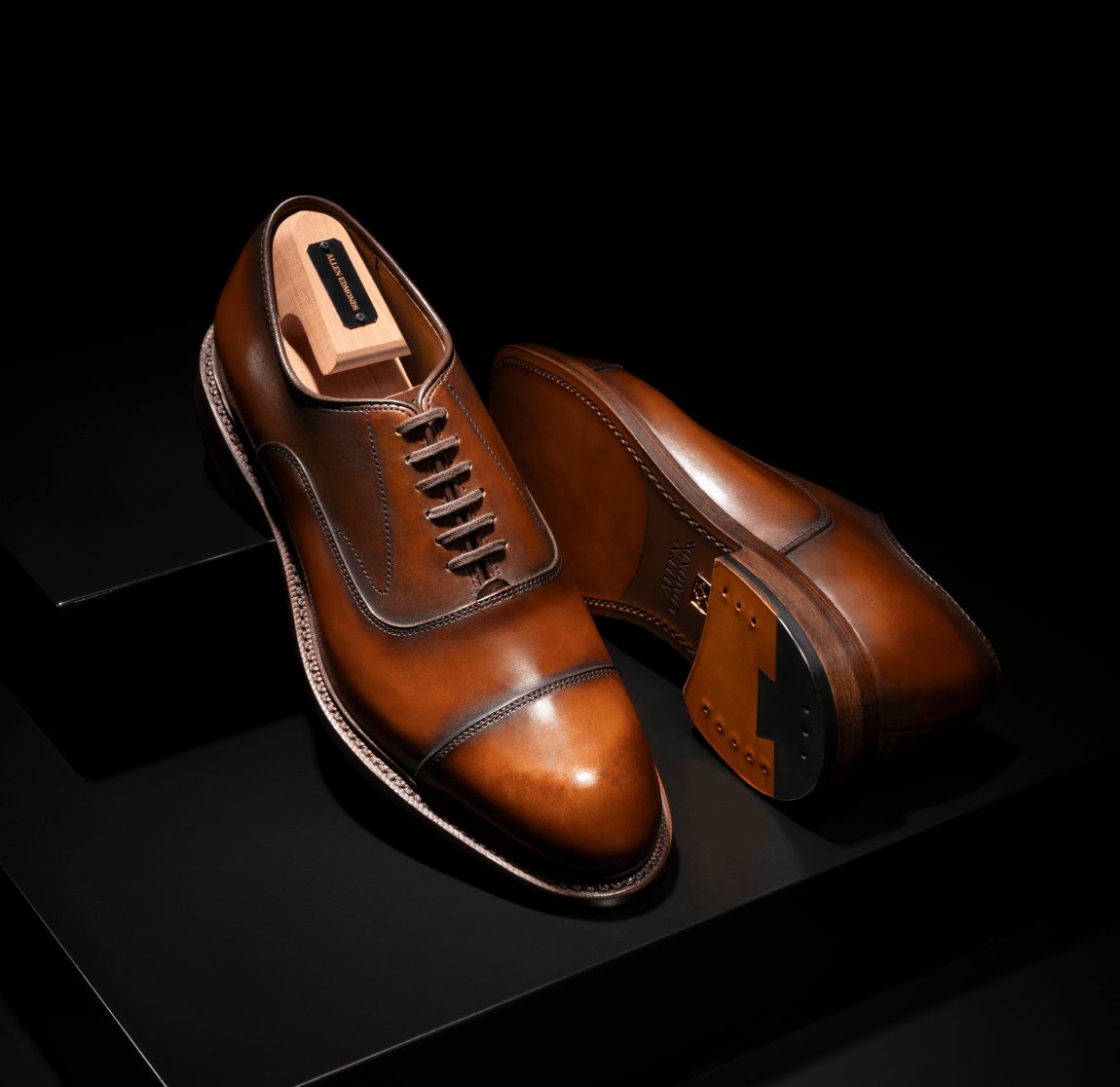 Brown leather Allen Edmonds Manhattan Cap-toe oxford dress shoes displayed with a branded cedar shoe tree against a dark background. 