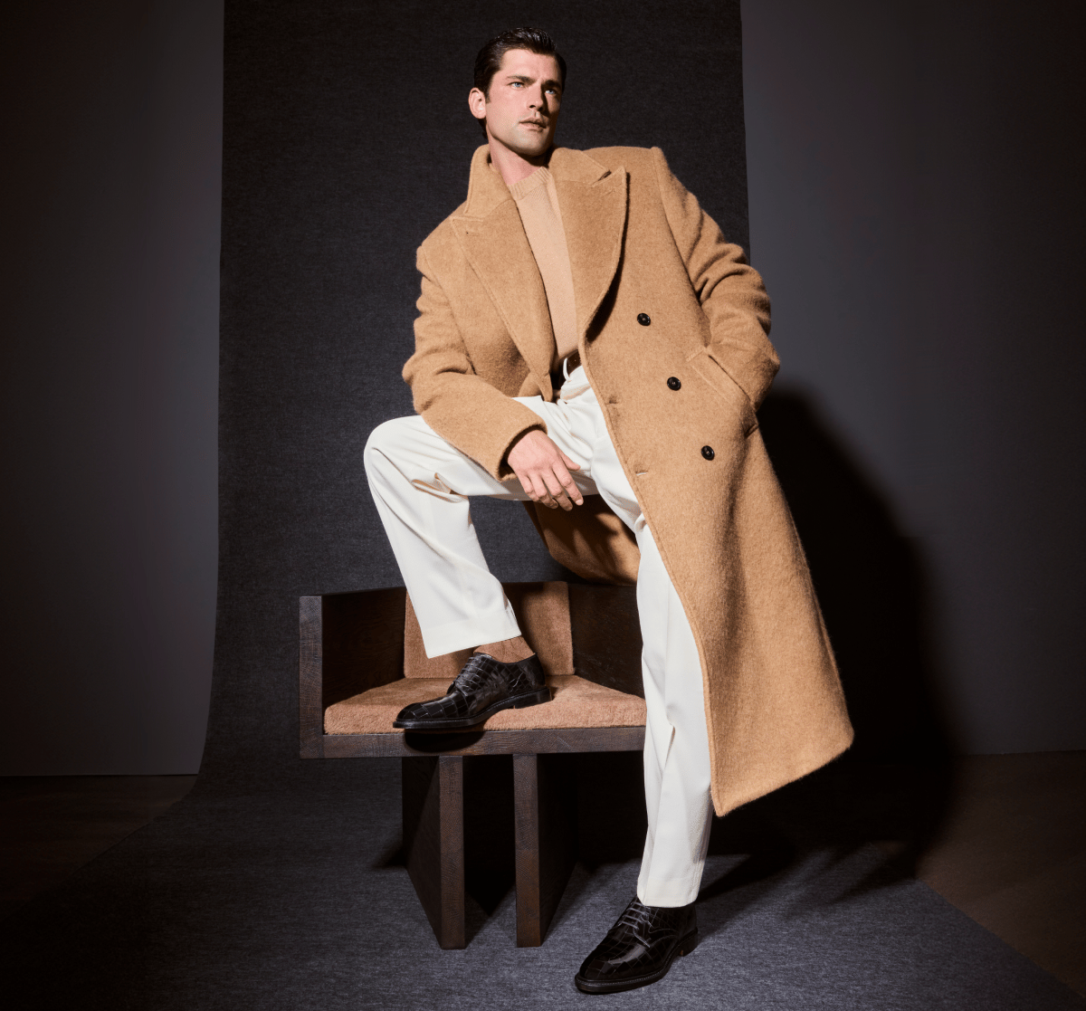 Black crocodile leather Allen Edmonds Maxwell Derby dress shoes and brown socks worn by a man in a tan overcoat and white trousers with one foot propped up on a wooden chair. 