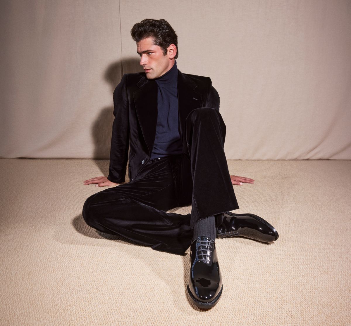 Black patent leather Allen Edmonds oxford dress shoes and dark socks worn by a man in a black velvet suit and navy turtleneck sitting on a neutral carpet 