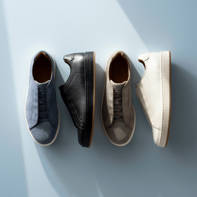 Top-down view of blue, black, grey, and white Allen Edmonds Banks stretch-lace sneakers with tan linings against a light blue background.