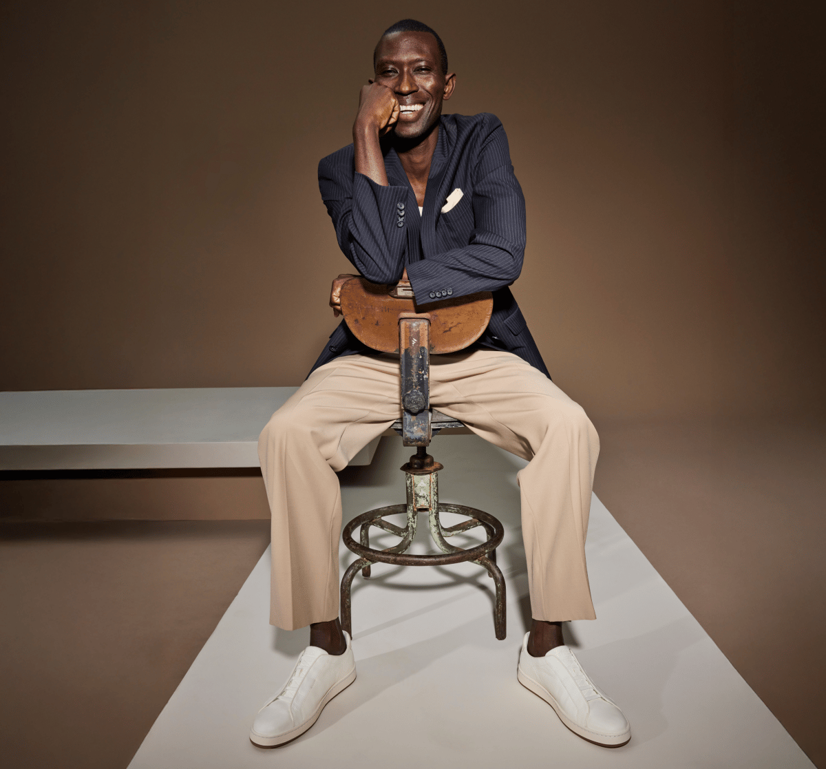 White leather Allen Edmonds Banks stretch-lace sneakers worn by a man in a navy pinstripe blazer and tan trousers sitting on a vintage metal stool