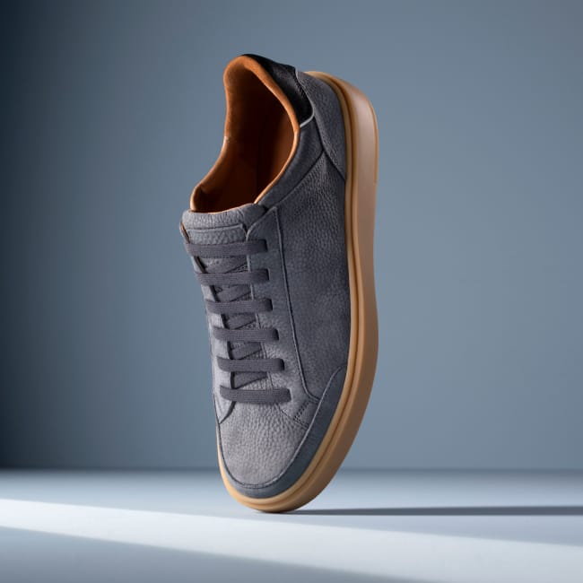 Grey leather Allen Edmonds Oliver stretch-lace sneaker with charcoal laces, tan lining, and a gum sole against a light blue background. 