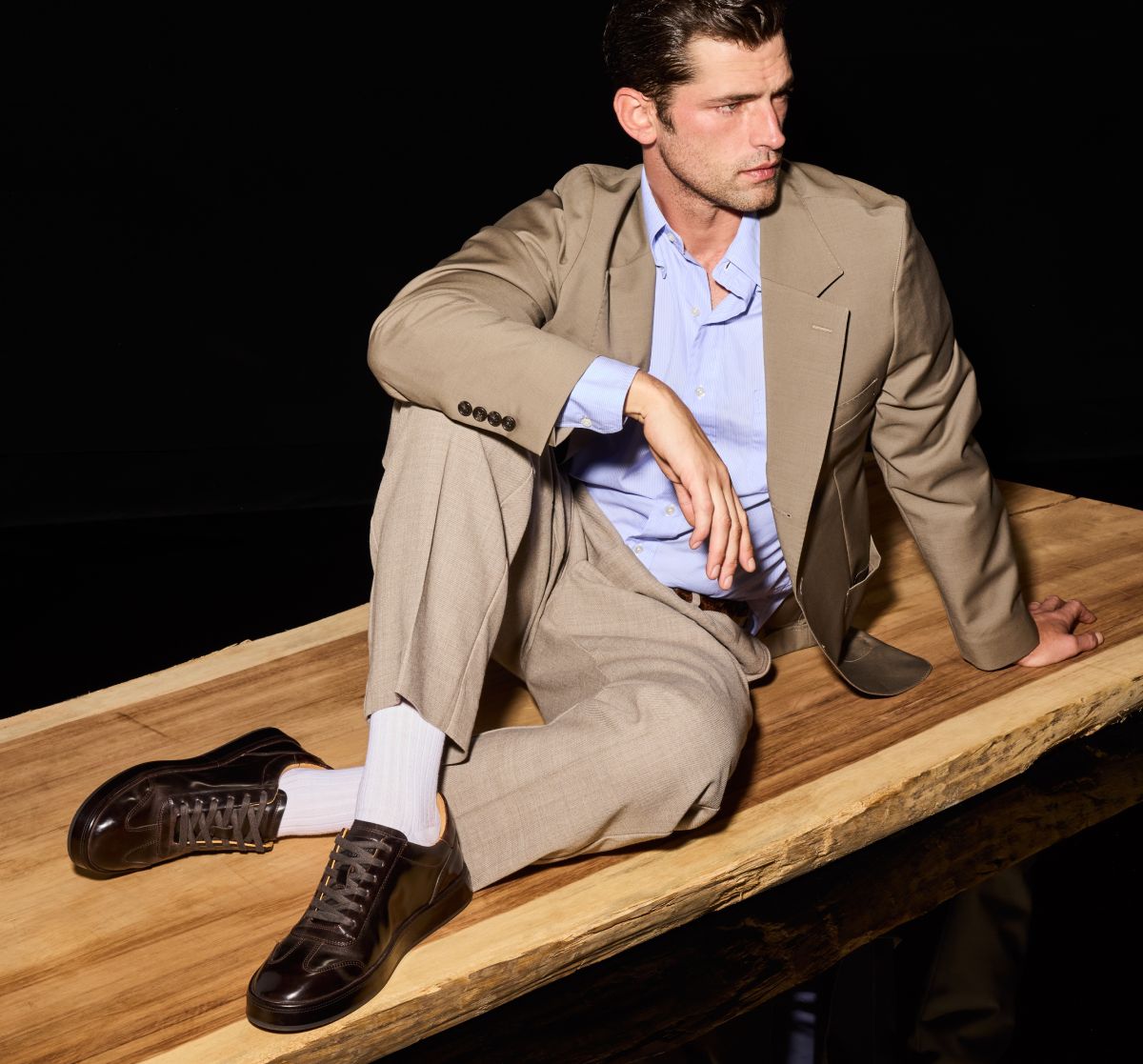 Dark brown Allen Edmonds Hawthorne sneakers and white socks worn by a man in a tan suit sitting on a wooden platform 